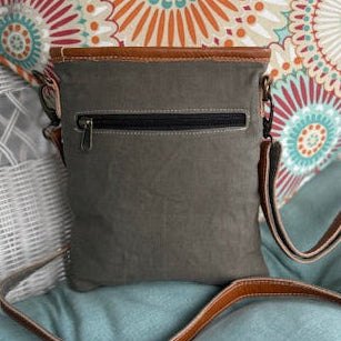 Gray crossbody bag with a colorful patterned pillow on a wicker chair.