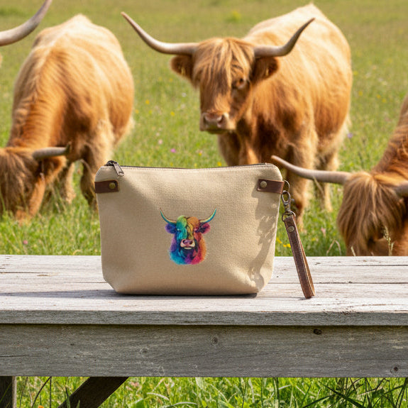 Highland cow multi-color print on a tan pouch or wristlet setting on a rustic table with Highland cows in the background.