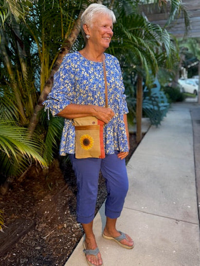 Woman in a blue floral top and pants wearing a tan recycled military canvas passport bag with a sunflower on the front