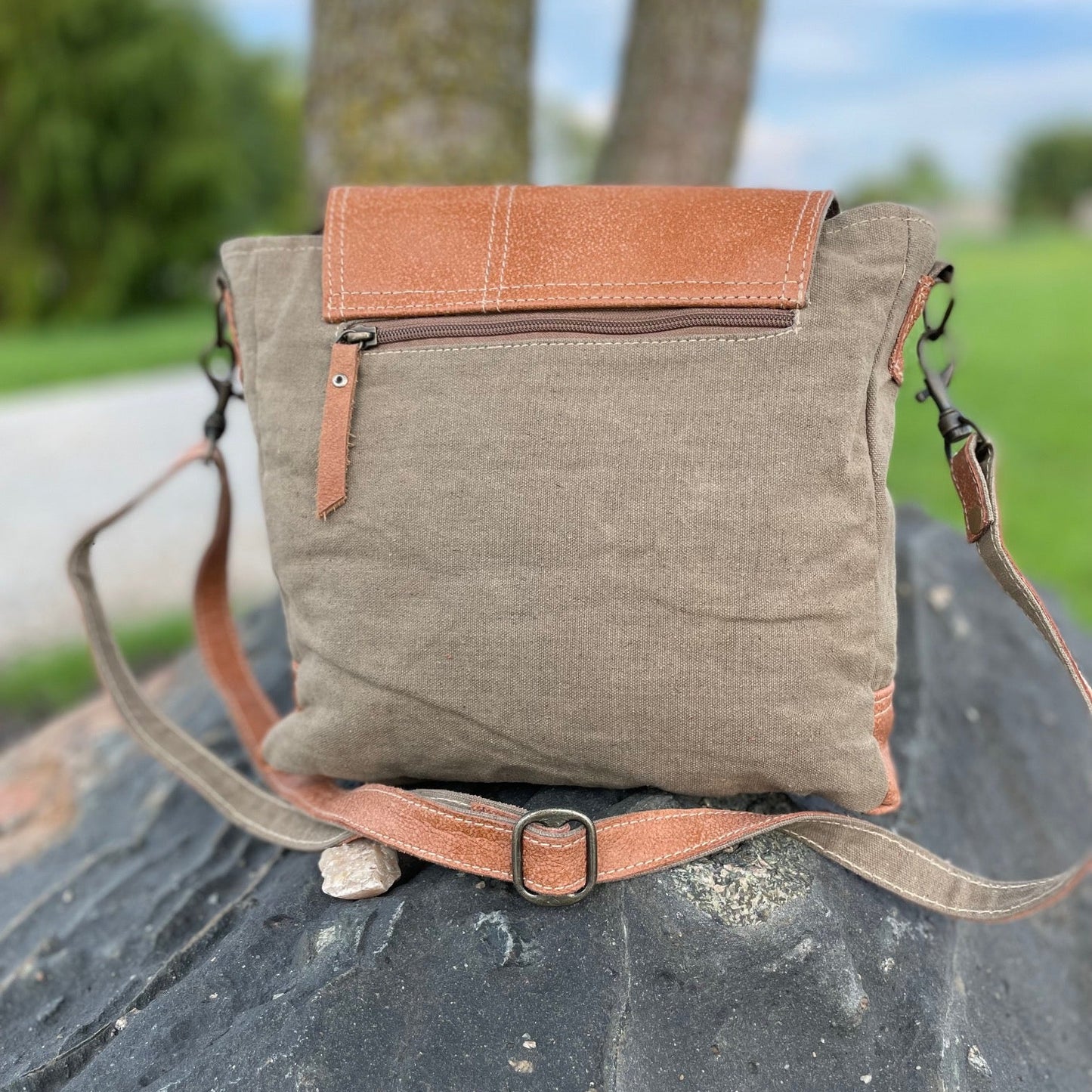 A Recycled Military canvas crossbody bag with tan leather trim and strap and front flap. Back view resting on a rock 