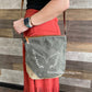 A woman wearing a Recycled Military Canvas  gray crossbody bag with white wings printed on the front and tan leather accent, standing against a wood-paneled wall.