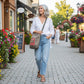 Tan recycled military canvas bag with a red label featuring a red Coffee label walking down a flower filled sidewalk in a small town being worn by a woman in jeans
