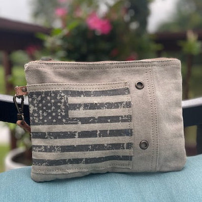 A recycled military canvas  wristlet zippered pouch with a distressed black-and-gray American flag print, resting on a light blue cushion.