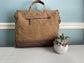 Brown leather bag on a light gray surface with a small plant in the foreground against a light blue wall.